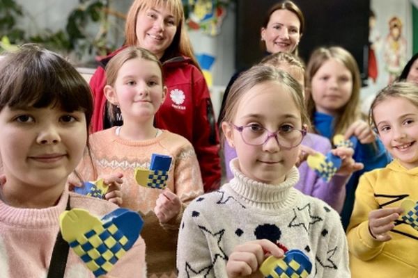 A group of children and adults smiling, holding colorful, woven paper hearts in a cheerful classroom setting.