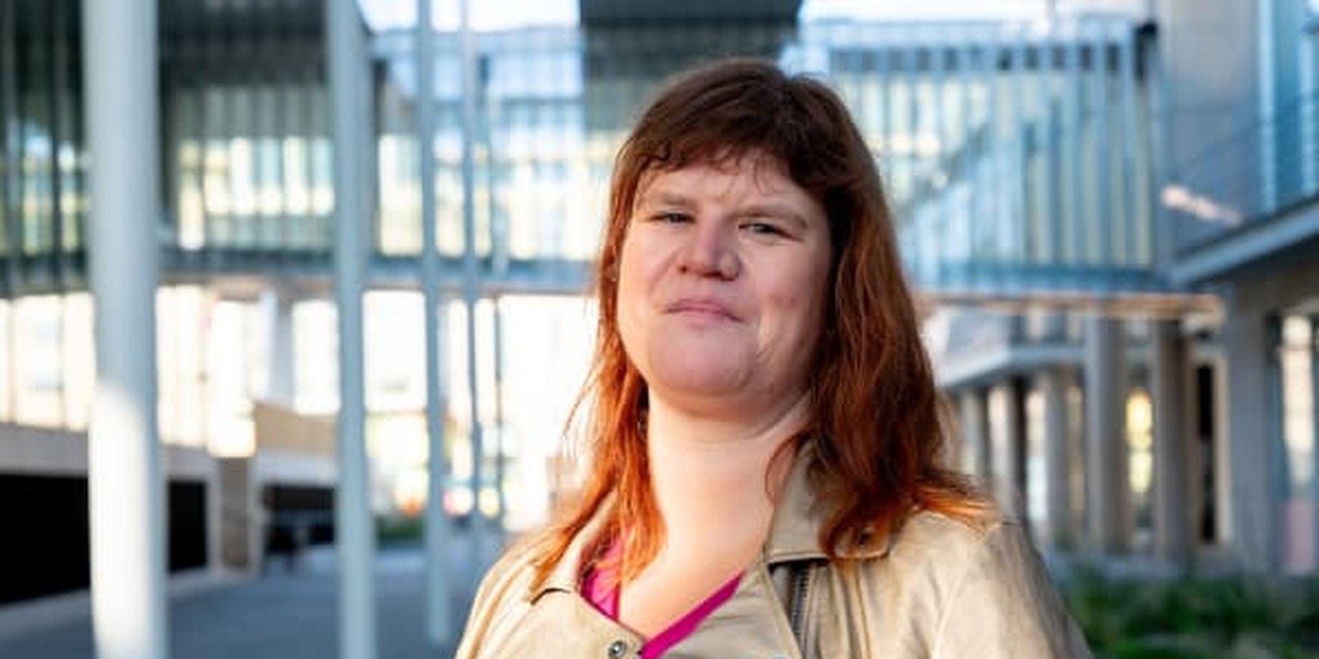 Person with brown hair wearing a beige jacket and pink top, standing outdoors in a modern architectural setting.