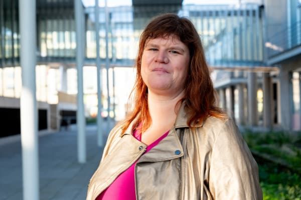 Person with brown hair wearing a beige jacket and pink top, standing outdoors in a modern architectural setting.
