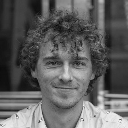 Smiling man with curly hair wearing a light blue shirt, standing in front of a blurred background.