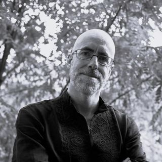 Bald man with glasses and a beard in a patterned shirt, standing outdoors with trees in the background, in black and white.