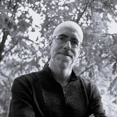 Bald man with glasses and a beard in a patterned shirt, standing outdoors with trees in the background, in black and white.