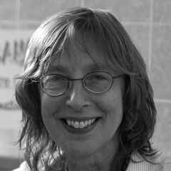 A smiling person with shoulder-length hair and glasses, wearing a light shirt, with a blurred, tiled background.