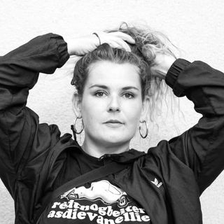 Person in a black jacket and hoop earrings, standing against a gray wall, tying their hair up with both hands.