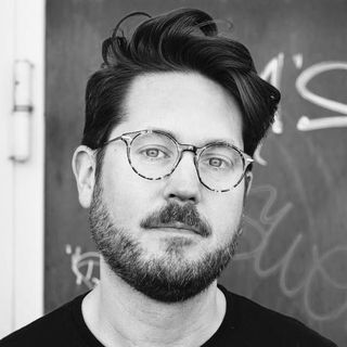 Man with glasses and a beard stands in front of a red wall with graffiti.