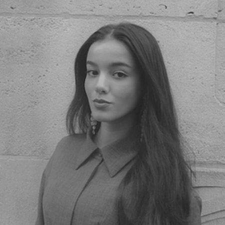 Black and white portrait of a woman with long hair and a serious expression, wearing a collared shirt, standing against a textured wall.