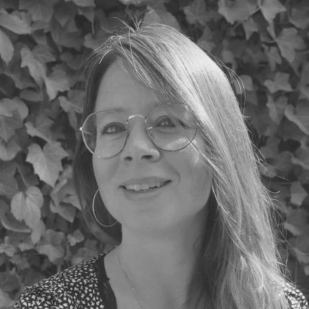 Smiling woman with glasses and long hair, wearing a patterned top, stands outside against a backdrop of ivy leaves. Black and white photo.