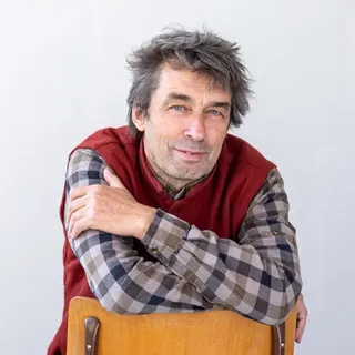 Man with tousled hair and plaid shirt, sitting backward on a wooden chair, arms crossed on the chair's back, against a plain white background.