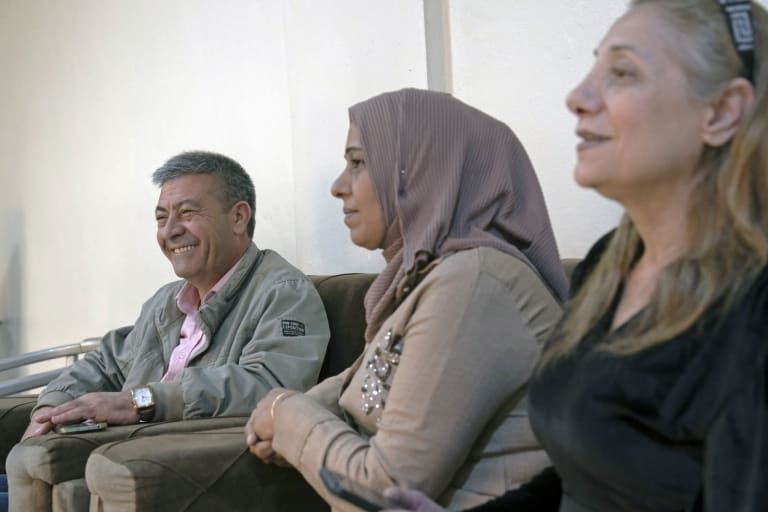 Three people sit together, smiling and talking. One man and two women, one in a hijab, are seated on a couch against a white wall.