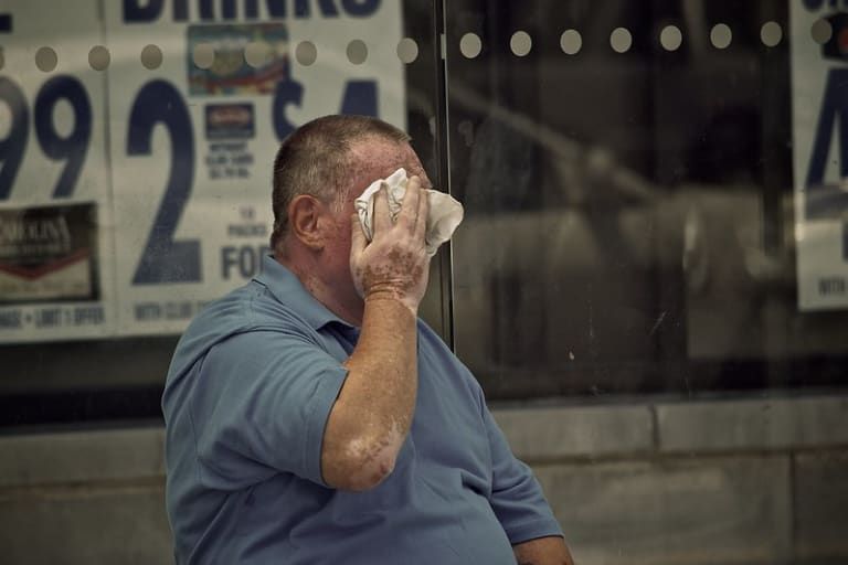 Man in blue shirt wipes sweat from his face with a cloth while sitting in front of a glass window displaying prices.