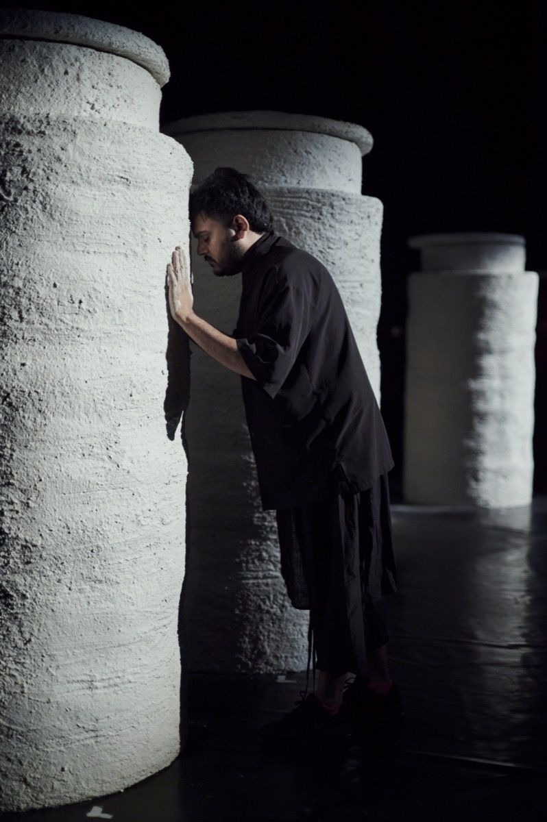 A person in dark clothing leans their forehead and hands against a large cylindrical column in a dimly lit setting.