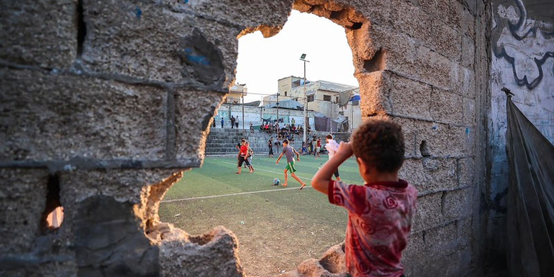 A young boy watches a soccer game through a large hole in a concrete wall, with players and spectators visible on the field in the background.