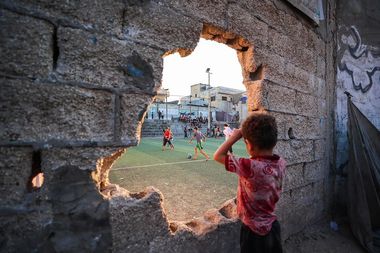 A young boy watches a soccer game through a large hole in a concrete wall, with players and spectators visible on the field in the background.