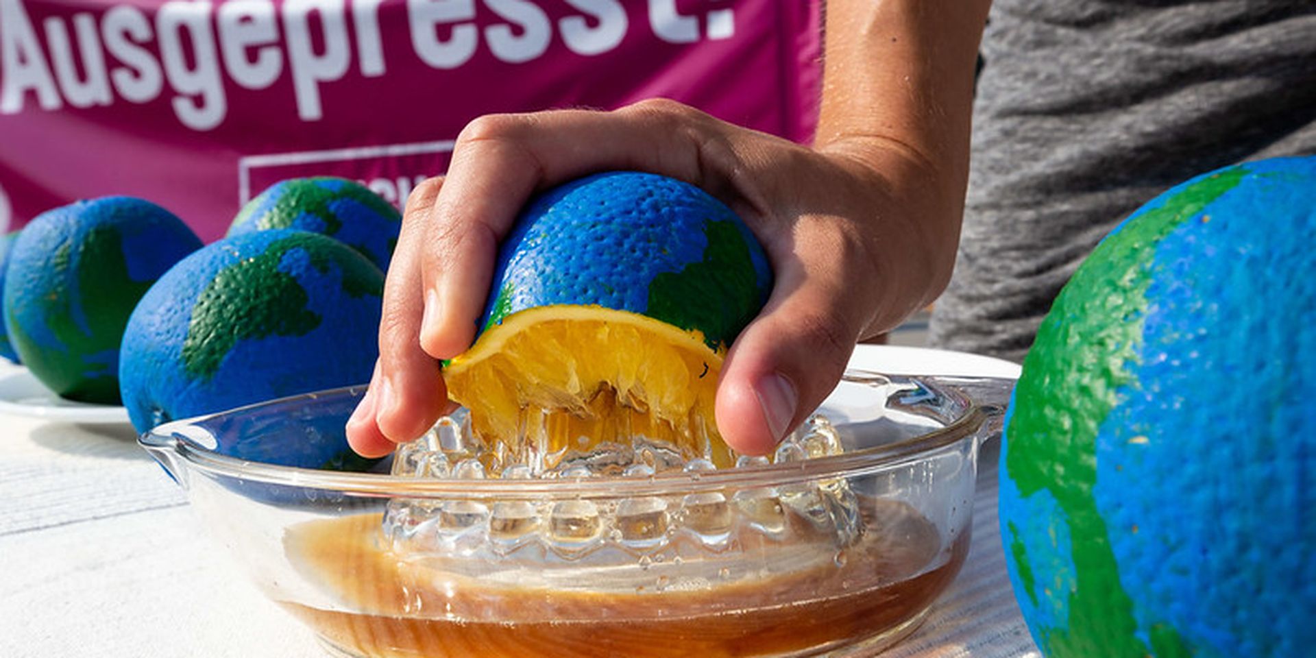 A hand squeezes a globe-shaped object resembling an orange into a glass dish. A banner with text is partially visible in the background.