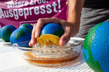 A hand squeezes a globe-shaped object resembling an orange into a glass dish. A banner with text is partially visible in the background.
