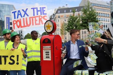 Protesters with signs saying "Tax the Polluters" and "Paris Agreement" confront a well-dressed man holding money by a gasoline pump.