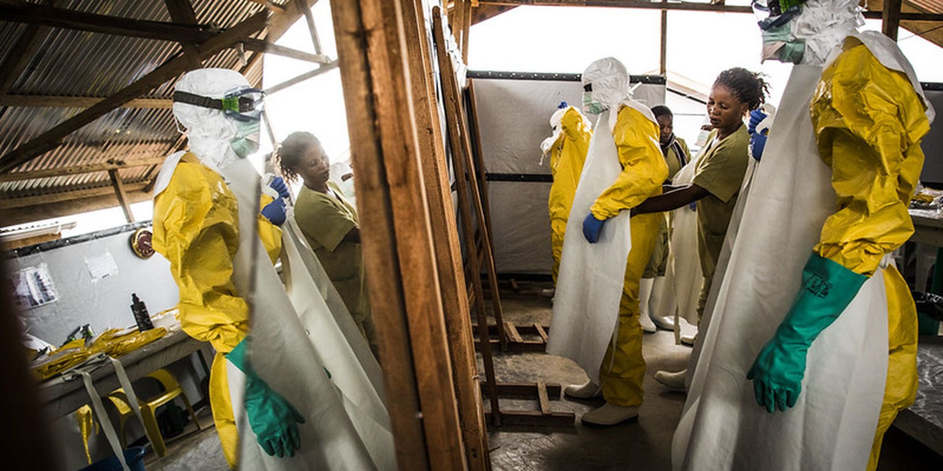 Healthcare workers in yellow protective suits and masks practice procedures in a wooden structure, with reflective surfaces on both sides.