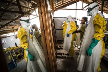 Healthcare workers in yellow protective suits and masks practice procedures in a wooden structure, with reflective surfaces on both sides.
