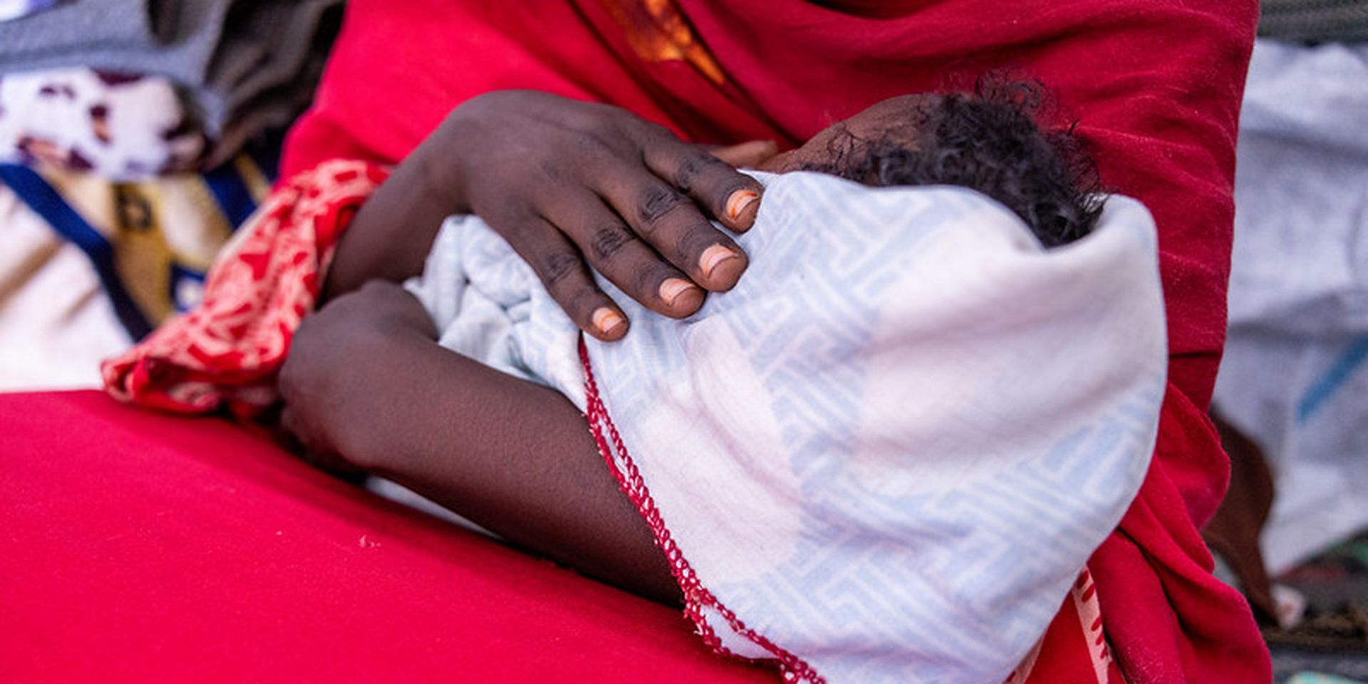 A close-up of a person in a red garment cradling an infant wrapped in a patterned white and red blanket.