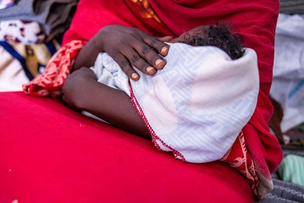A close-up of a person in a red garment cradling an infant wrapped in a patterned white and red blanket.