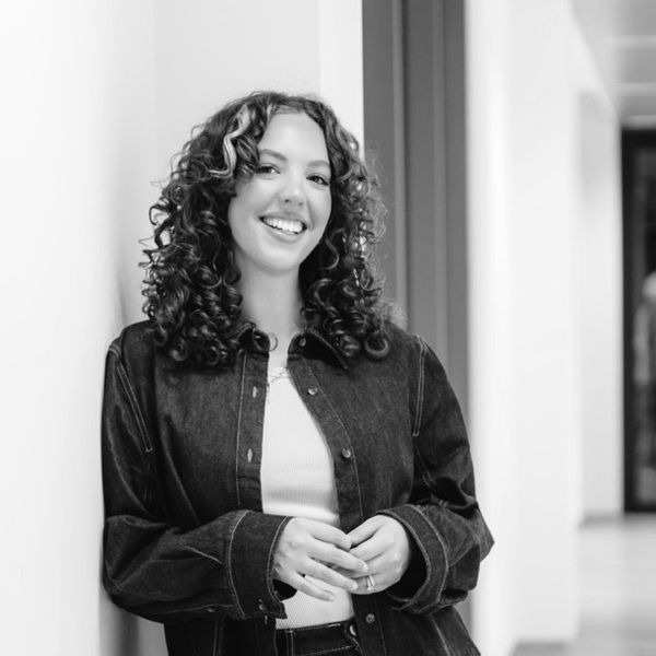 Smiling woman with curly hair leans against a wall in an office hallway, wearing a denim outfit and a white top.