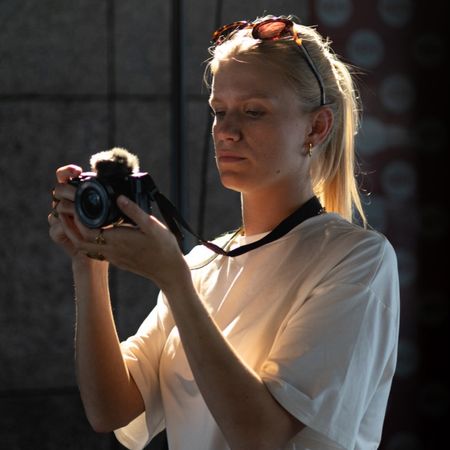 Woman with sunglasses on head examines a camera, standing in a softly lit room with dark walls and patterned wallpaper in the background.
