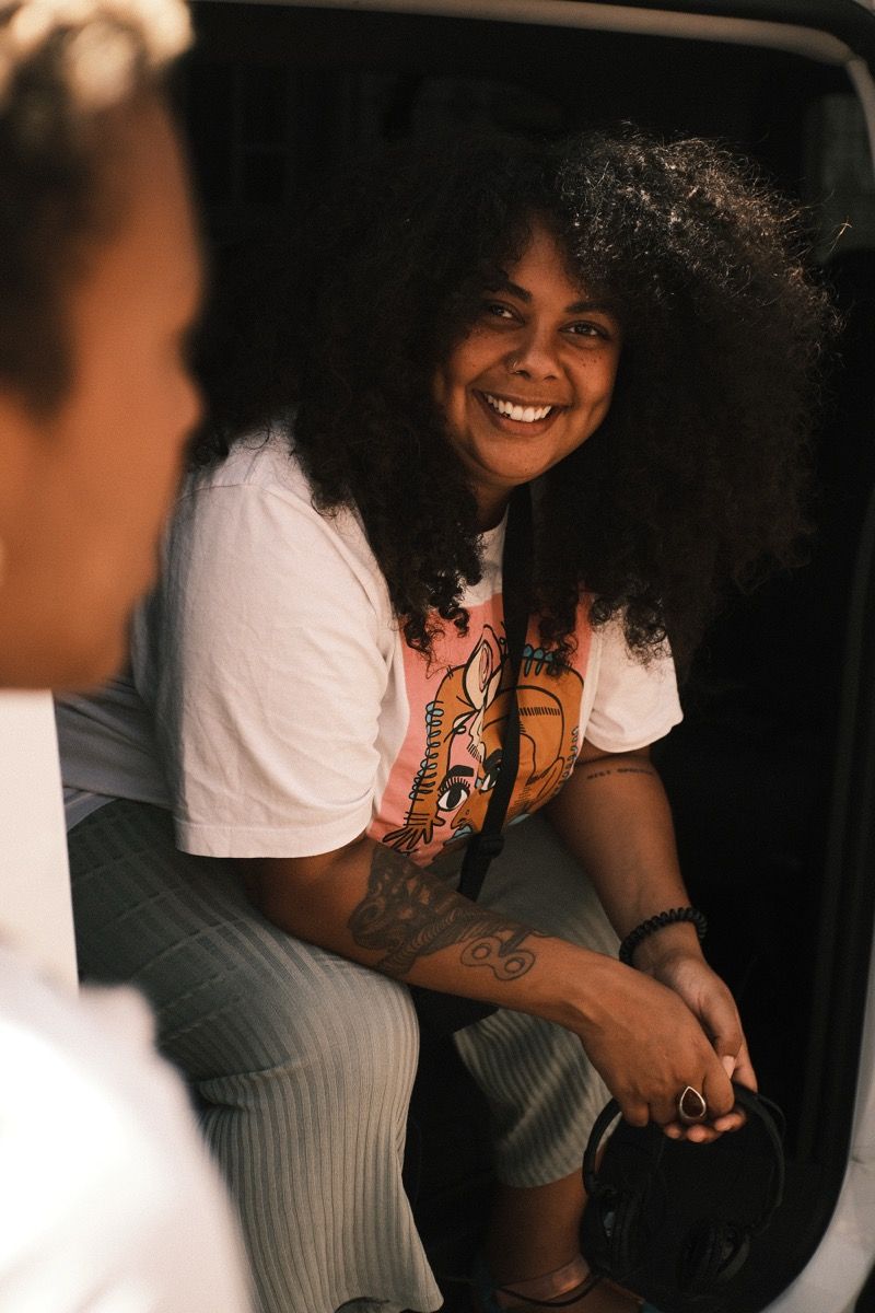 Smiling person with curly hair and tattoos, sitting in a car holding headphones, wearing a graphic t-shirt and striped pants.
