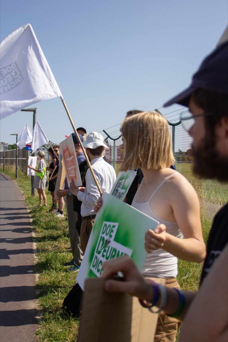 People holding signs and flags stand in a line during a sunny protest along a sidewalk next to a fence.