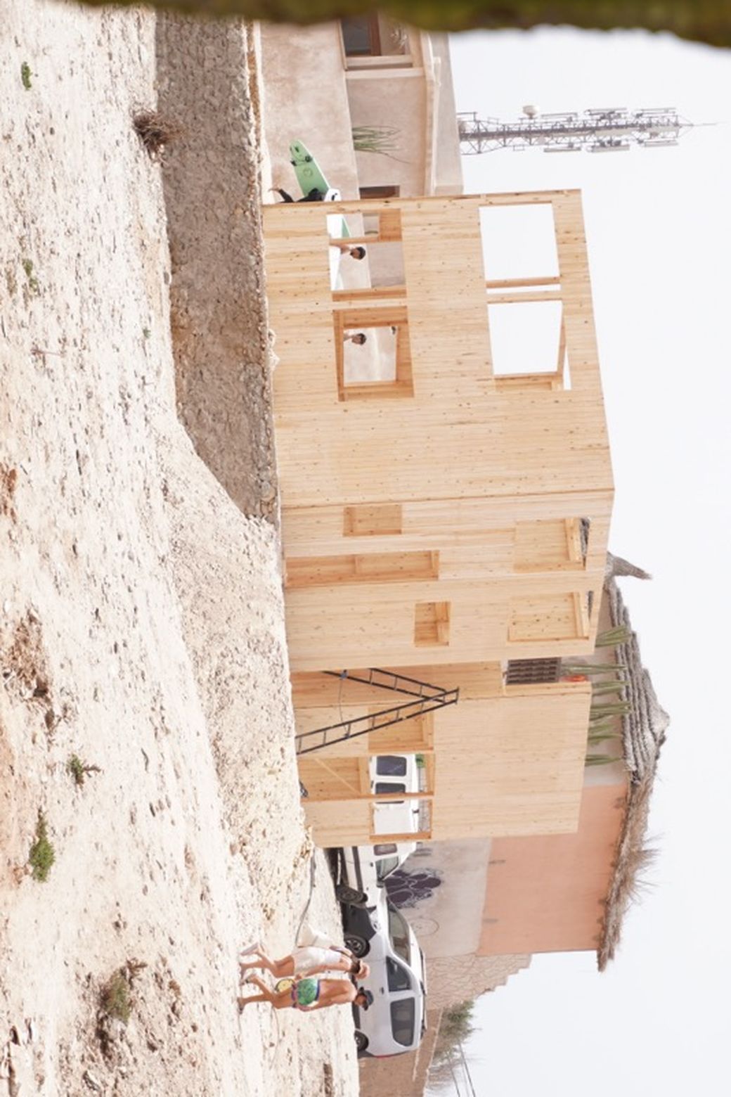 Wooden house under construction on a sandy terrain, with two people walking nearby and cars parked in the background.