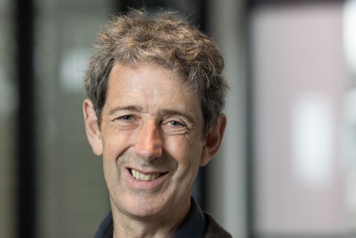 Smiling man with gray hair wearing a brown jacket and blue shirt, standing indoors with a blurred background.