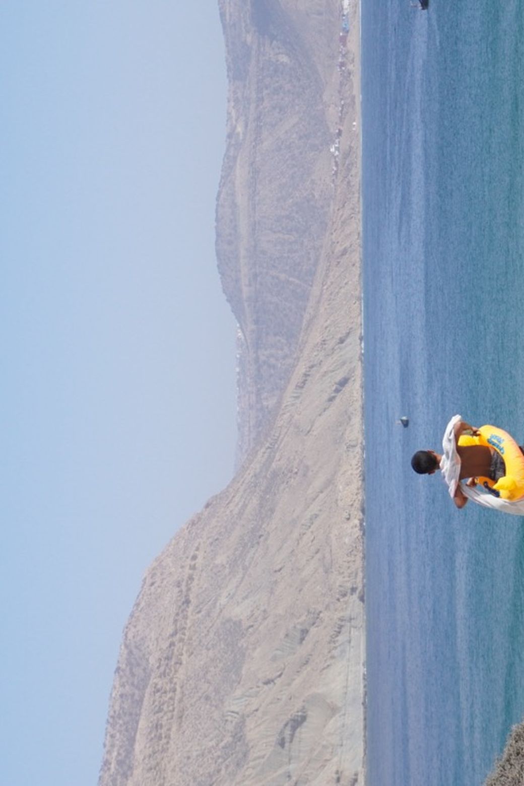 Child with a yellow floatie stands on a sandy hill overlooking a blue ocean with gentle waves and distant mountains under a clear sky.