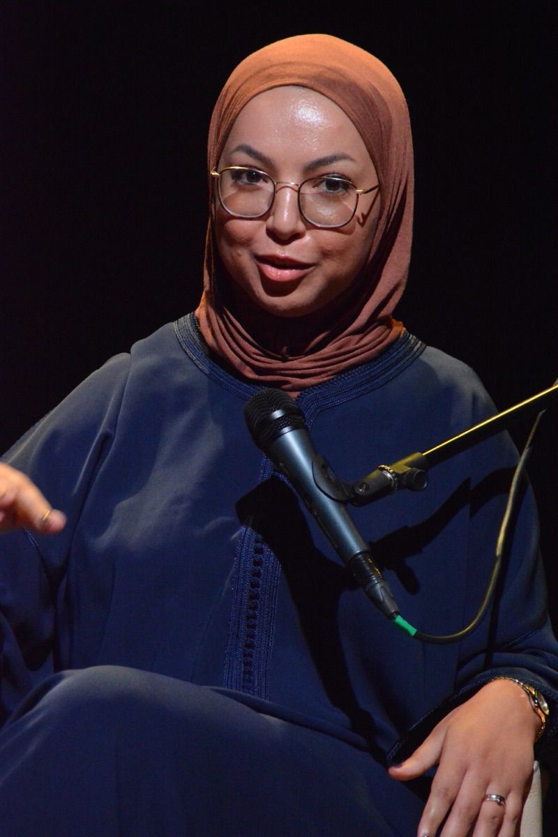 A woman in a brown hijab and glasses speaks into a microphone, wearing a dark outfit against a black background.
