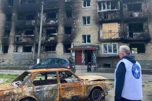 Een hulpverlener van Dokters van de Wereld staat naast voor een door raketten beschadigd gebouw naast een uitgebrande auto