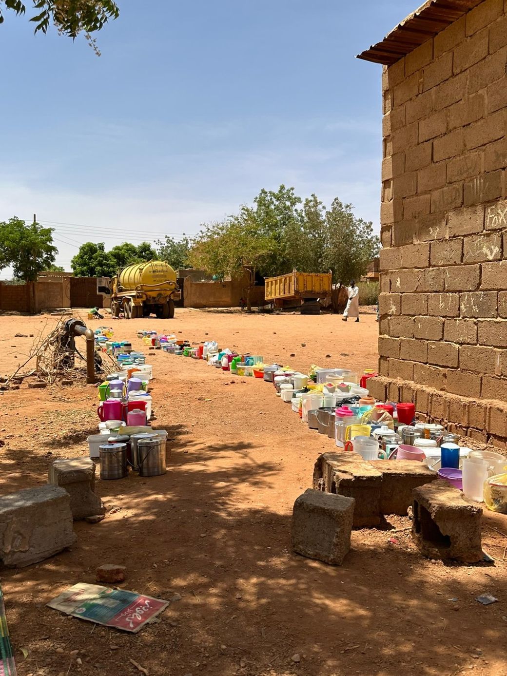Colorful containers line up on a dirt path near a brick building under a sunny sky, with water trucks and trees in the background.