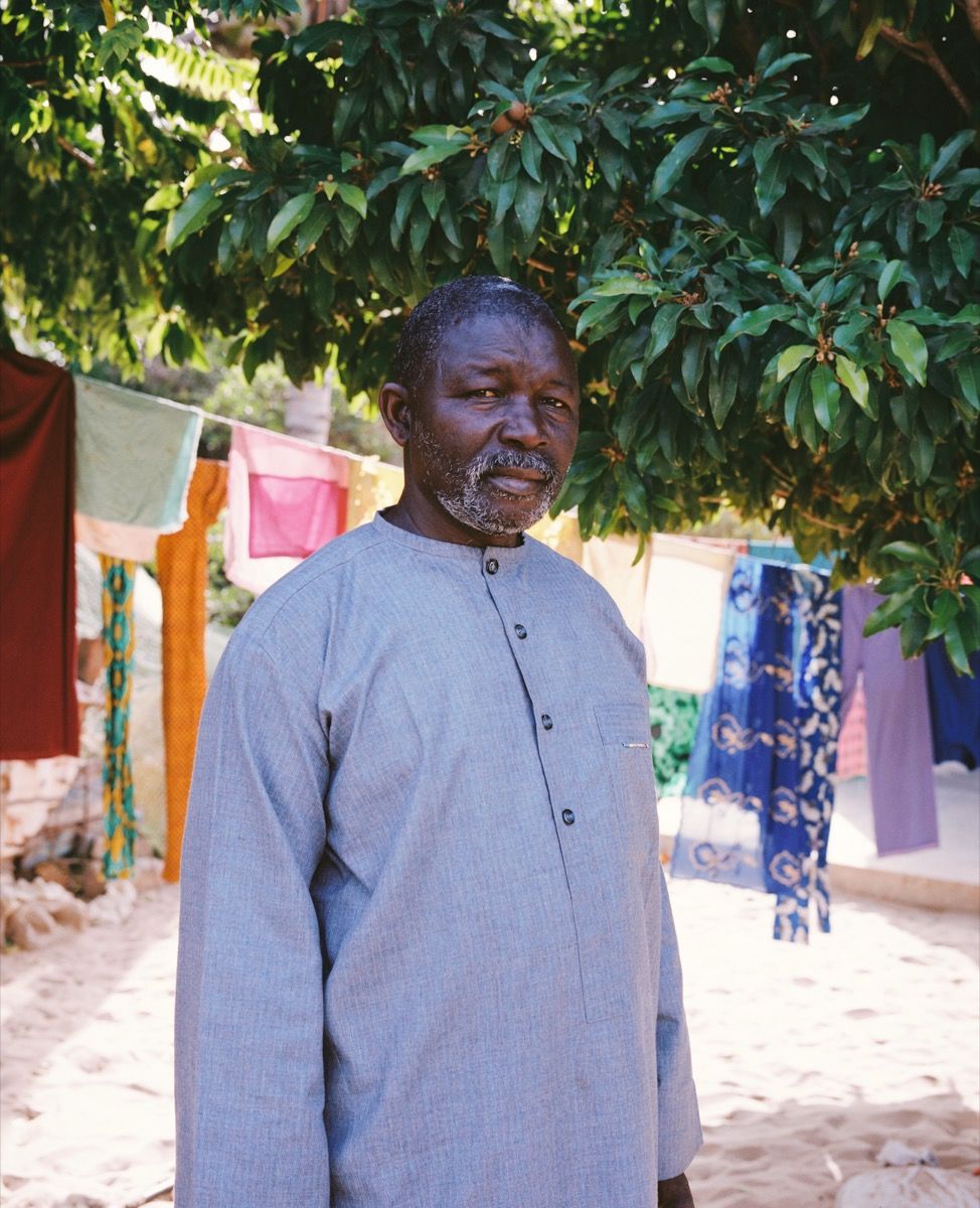 A man in a blue shirt stands under a leafy tree, with colorful fabrics hanging in the background.