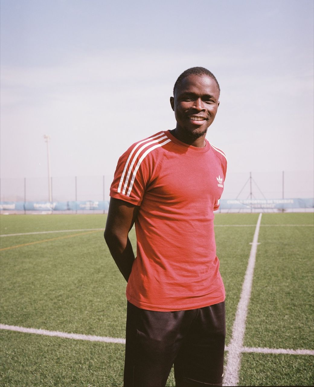 Smiling man in red shirt stands on green sports field, hands behind back, with clear sky in the background.