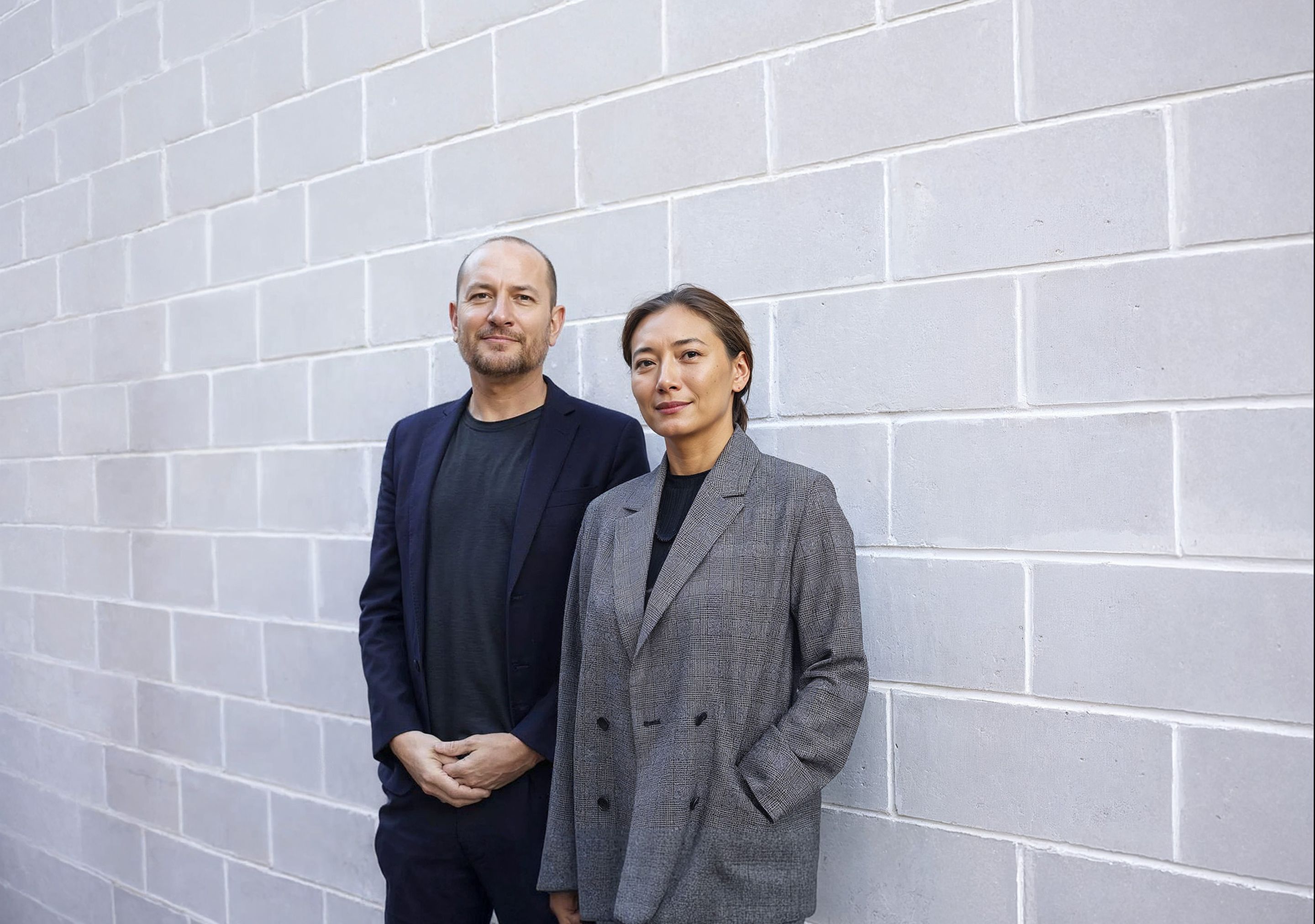 Two people in business attire stand against a light gray brick wall, looking at the camera.