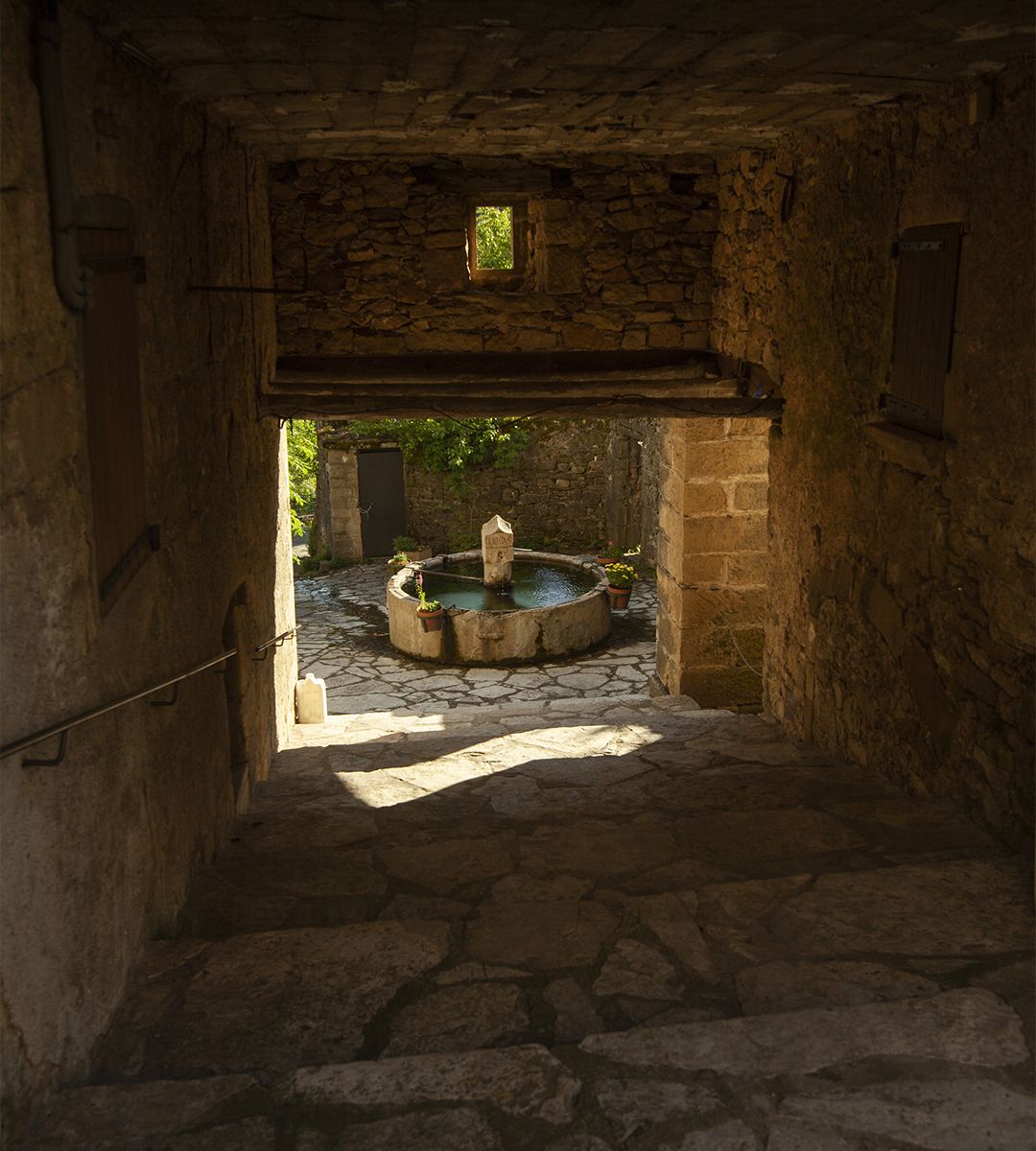Fontaine en pierre au cœur d’un village ancien cévenol vue depuis une ruelle voûtée | Atelier Tuffery