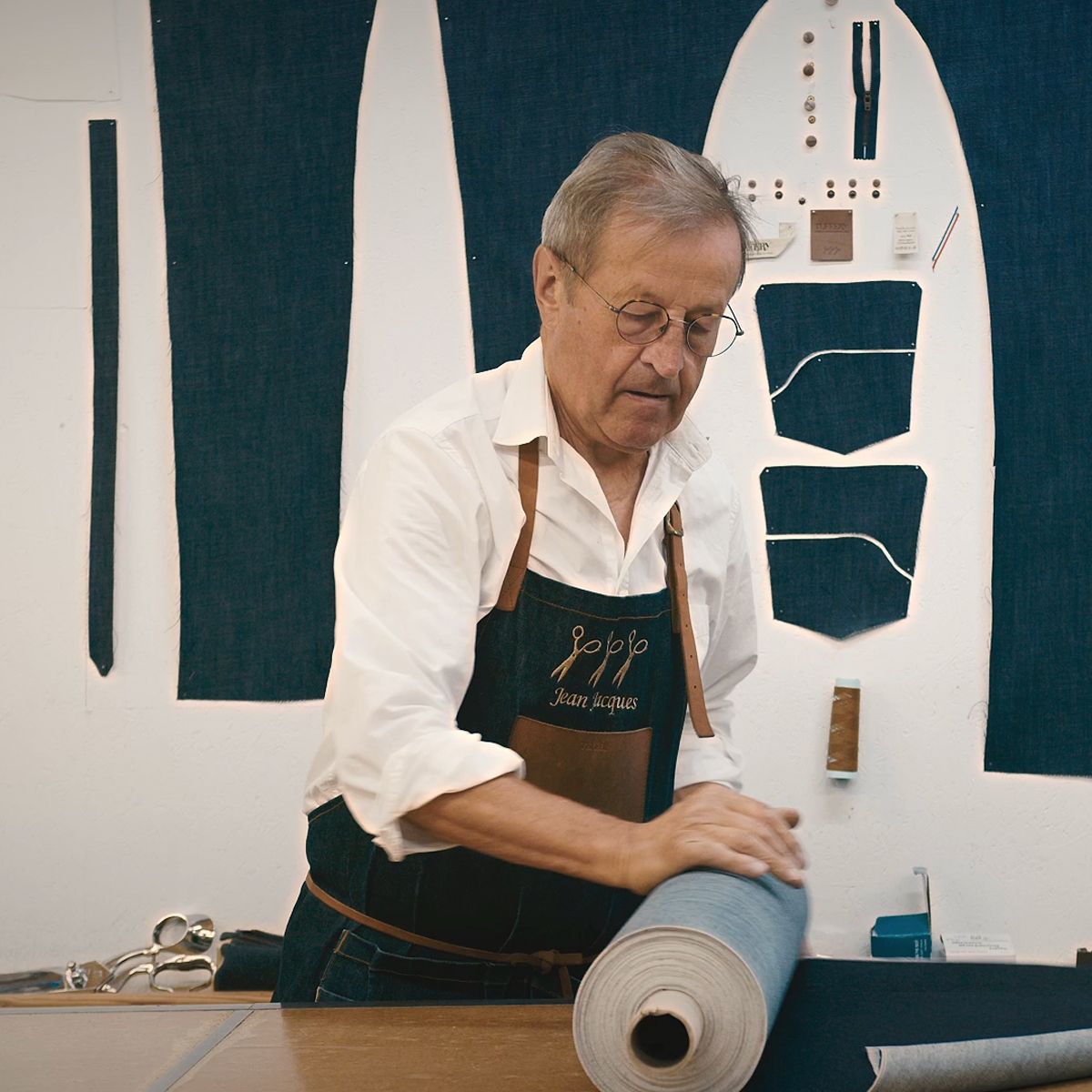 Un tailleur, Jean-Jacques Tuffery, en tablier déroule un tissu sur une table, avec des patrons de couture et des outils accrochés au mur derrière lui. | Atelier Tuffery