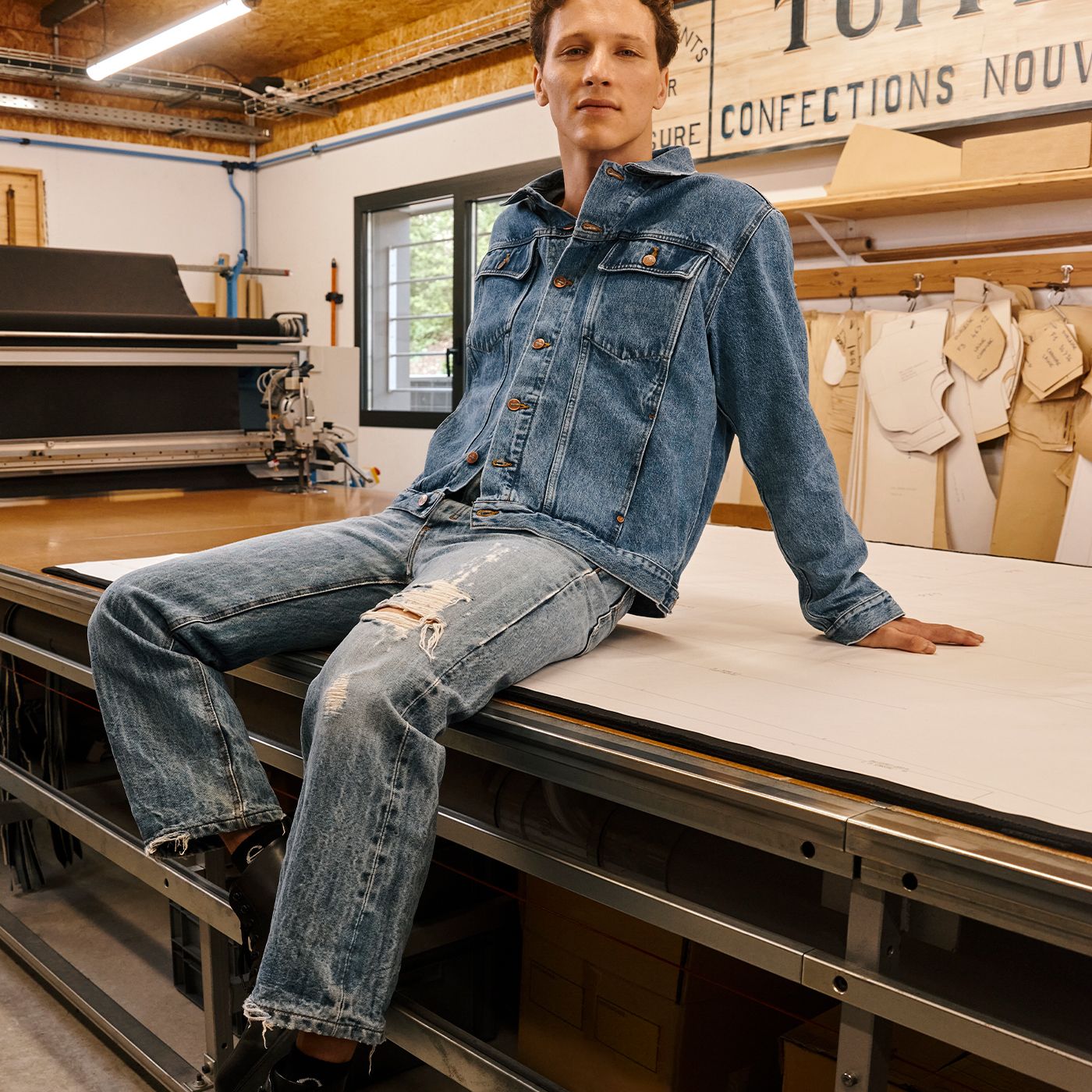 Homme portant une veste et un jean en denim assis sur une grande table. | Atelier Tuffery