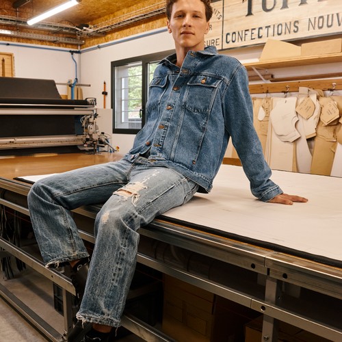 Homme portant une veste et un jean en denim assis sur une grande table. - Atelier TUFFERY