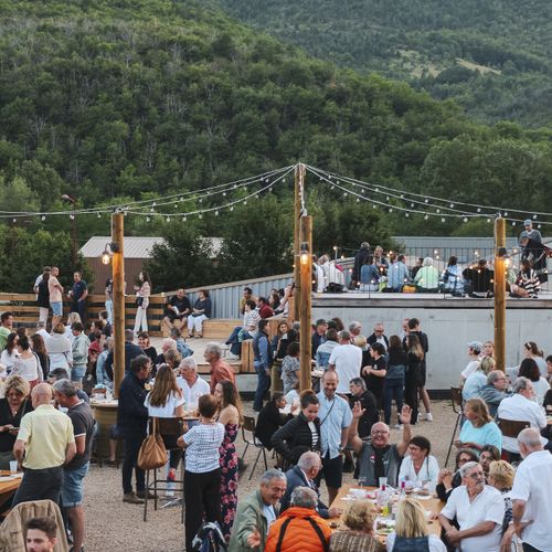 Soirée Les Heures Bleues sur les toits de la manufacture, avec de nombreuses personnes réunies autour de tables en bois, dans une ambiance conviviale et musicale. - Atelier Tuffery
