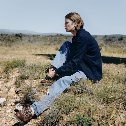 Homme assis dans un paysage sec, portant une veste en laine et un jean, pour évoquer le passage de la laine au vêtement. - Atelier Tuffery