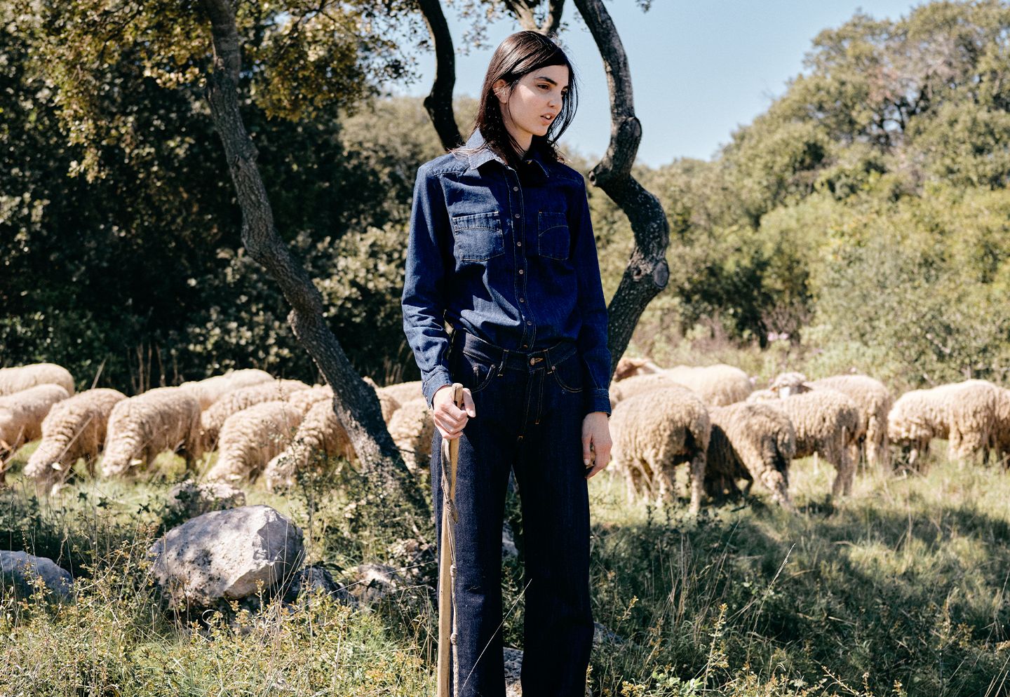 Femme portant une tenue en denim dans un paysage cévenol, avec un troupeau de brebis en arrière-plan. | Atelier Tuffery