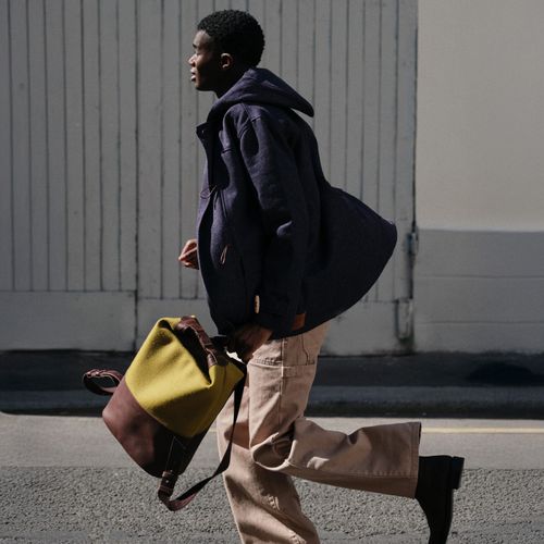 Un homme qui court avec un manteau, un cargo écru et un sac à dos en laine et cuir - Atelier Tuffery