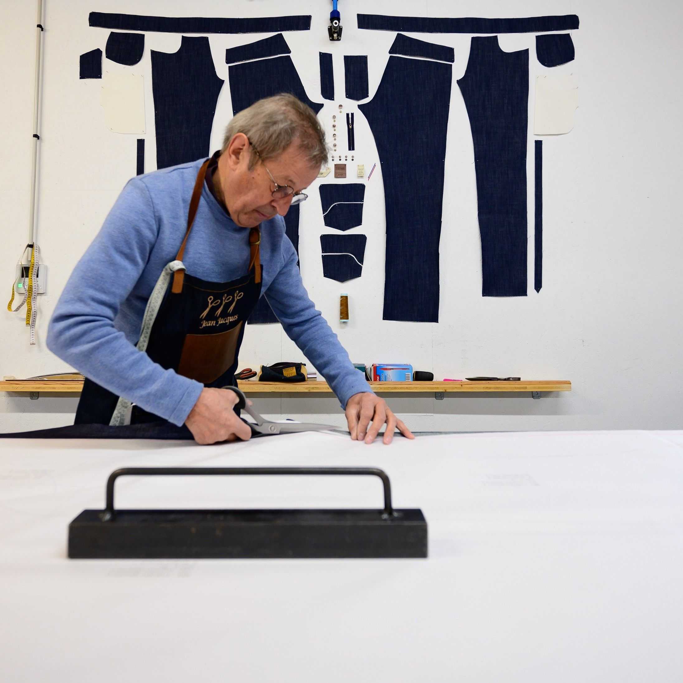 Un tailleur, Jean-Jacques Tuffery, en tablier déroule un tissu sur une table, avec des patrons de couture et des outils accrochés au mur derrière lui. | Atelier Tuffery