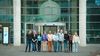 A group of eleven people stand in front of a glass building entrance with a large circular logo.