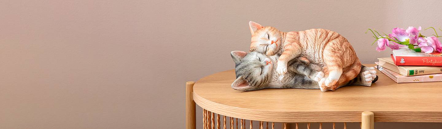 Zwei realistische Katzenfiguren, eine orangefarbene und eine graue, kuscheln auf einem Holztisch neben Büchern und rosa Blumen.