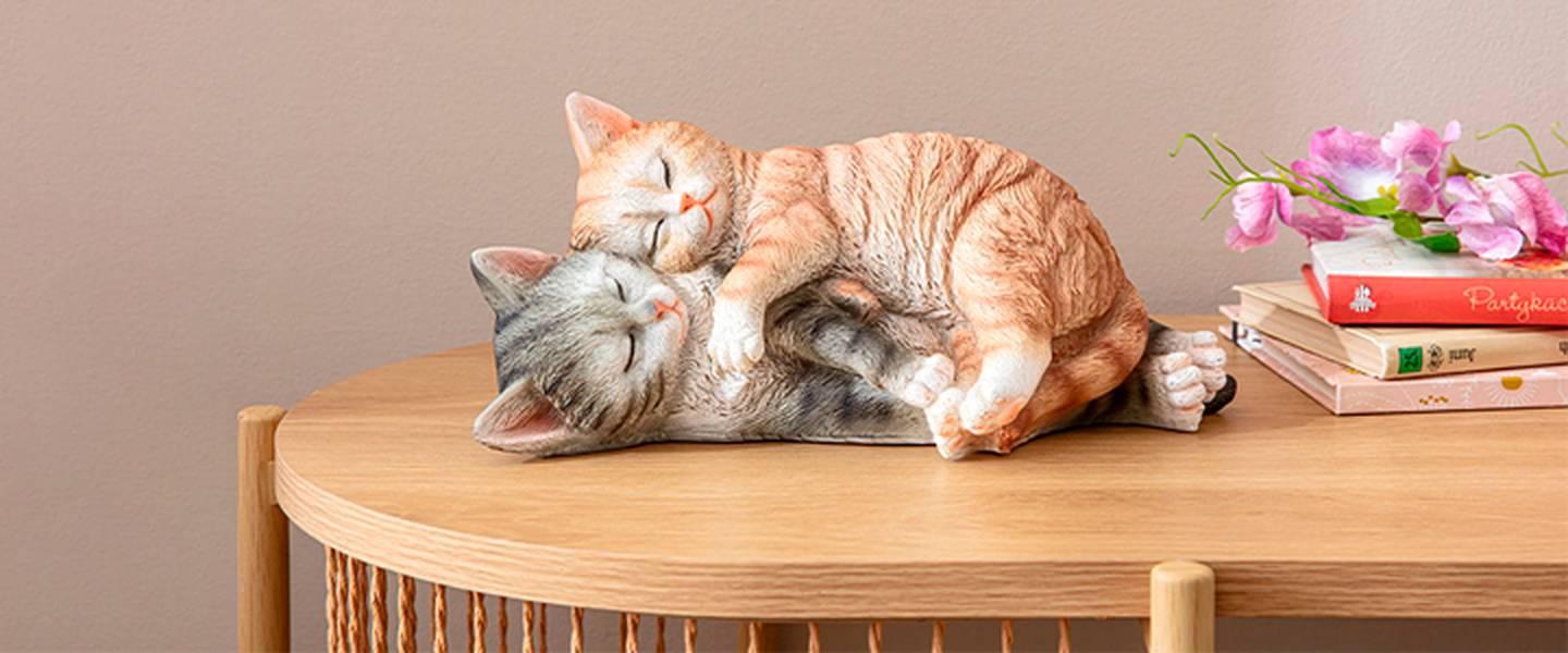 Zwei realistische Katzenfiguren, eine orangefarbene und eine graue, kuscheln auf einem Holztisch neben Büchern und rosa Blumen.