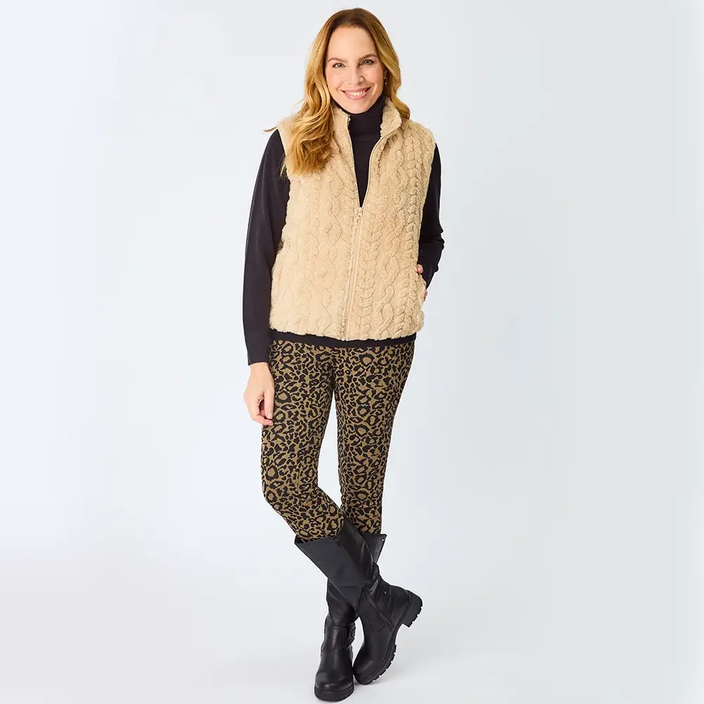 Frau in einer beigen Weste und einer Hose mit Leopardenmuster, die schwarze Stiefel und einen schwarzen Rollkragenpullover trägt, steht lächelnd vor einem weißen Hintergrund.